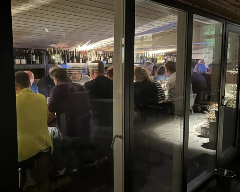 Folk sidder tæt sammen ved langbord i hyggeligt vinlokale med flasker på hylderne, set gennem glasdør om aftenen.